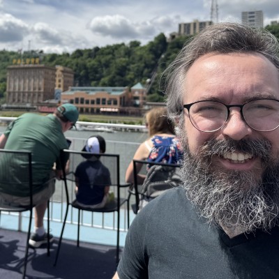 Close-up selfie of a smiling man with round glasses, a full graying beard, and a dark shirt on the outdoor deck of a boat. In the background, other passengers sit in chairs looking out toward a riverfront city skyline with buildings and a lush, tree-lined hill under a partly cloudy sky.